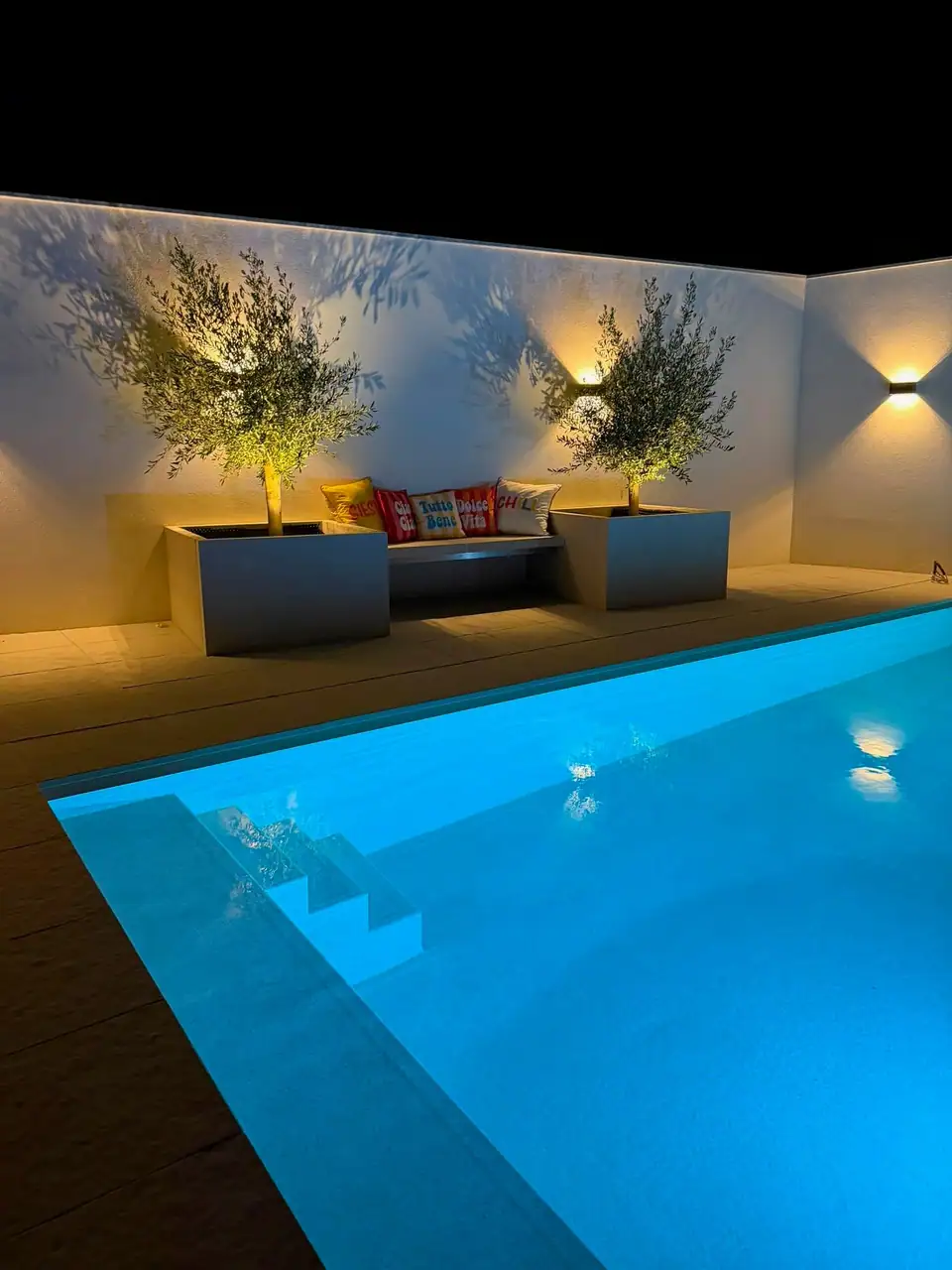 Beleuchteter Pool mit Terrassenplatten und Hochbeeten bei Nacht